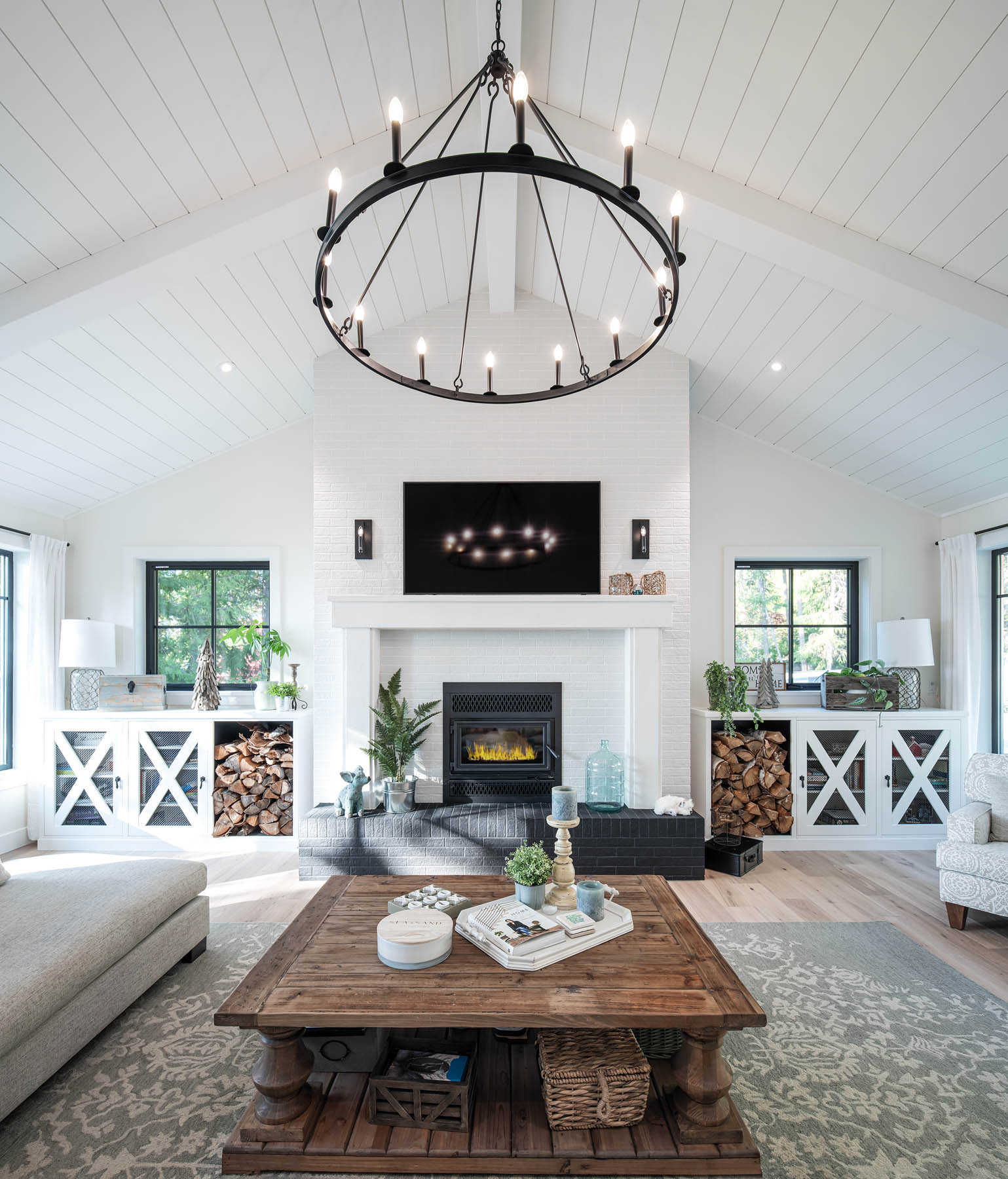 A bright and airy living room featuring a vaulted ceiling with white beams, an elegant chandelier, a cozy fireplace with a stocked woodpile, flanked by built-in shelving, and stylish, comfortable furnishings.