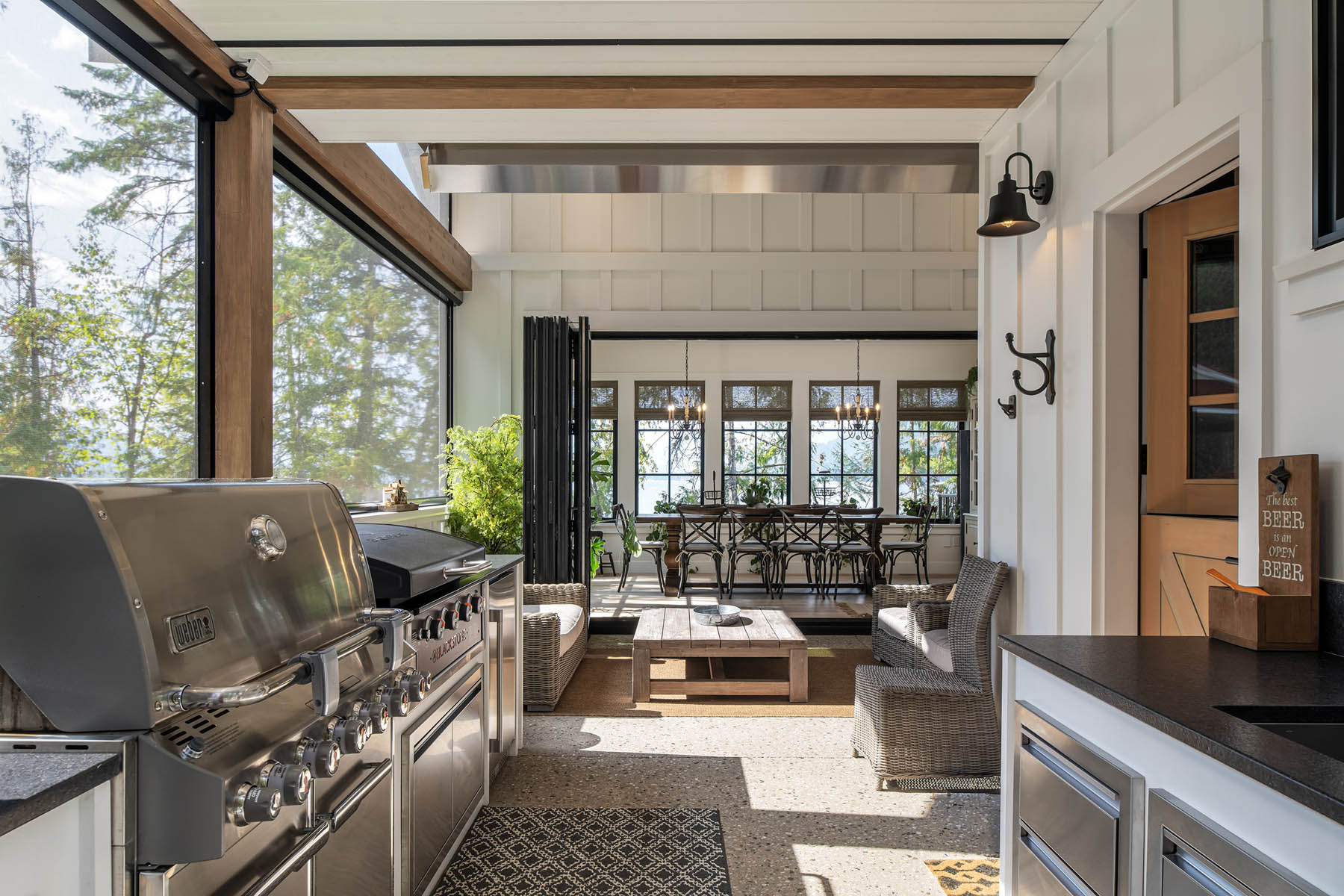An elegant indoor-outdoor living space equipped with a modern grill kitchen area, featuring an open wall leading to a comfortable patio with a dining set, all bathed in natural light and surrounded by lush greenery.