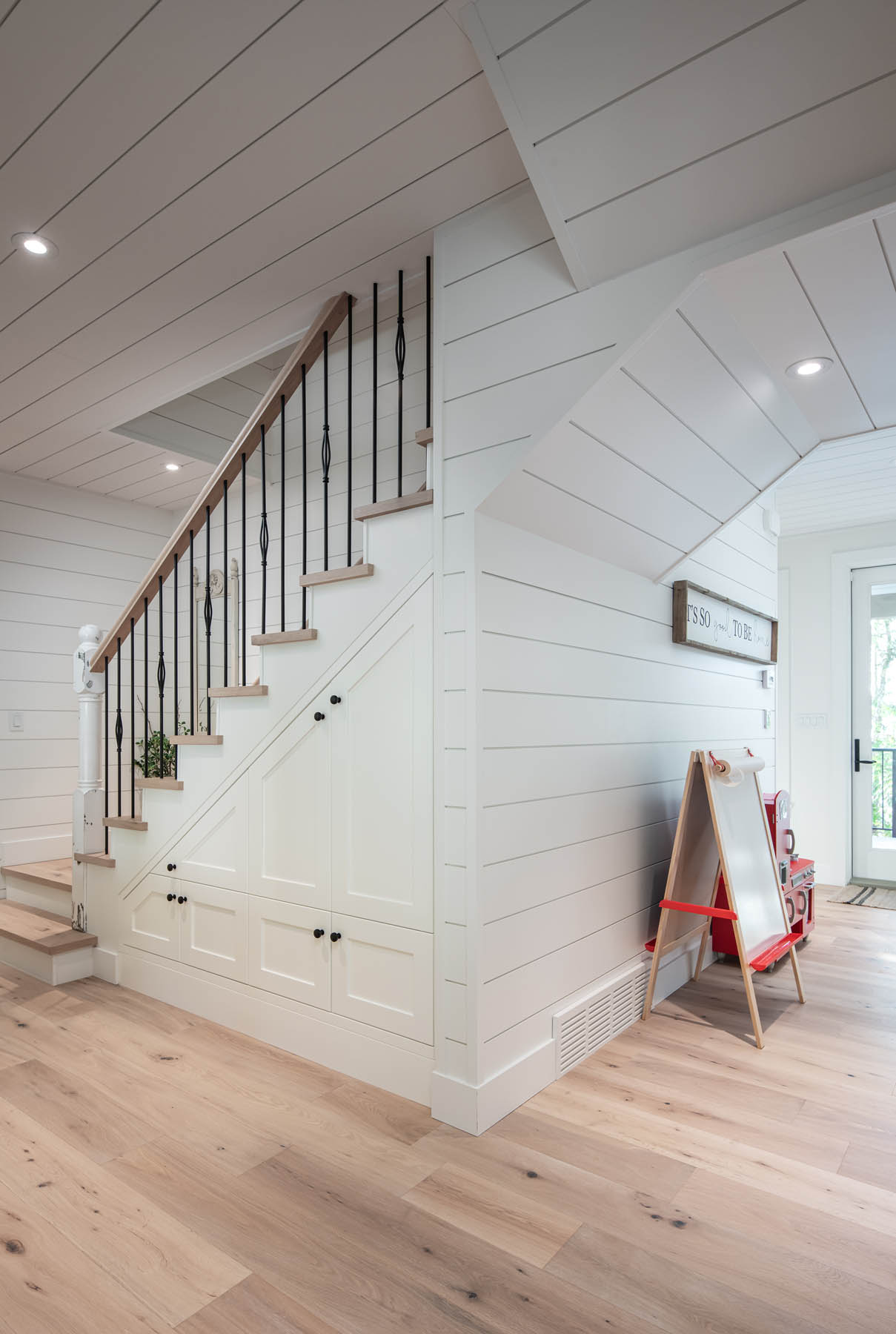 A bright and airy entryway featuring a wood staircase with black metal balusters, white shiplap walls, and built-in storage beneath the stairs.