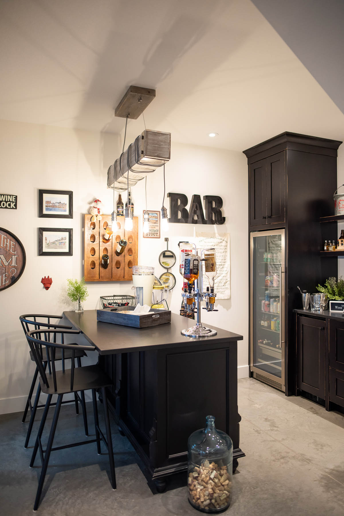 A cozy and stylish home bar corner featuring a black bar counter with stools, a wine rack on the wall, and a glass-fronted fridge, all under warm pendant lighting.